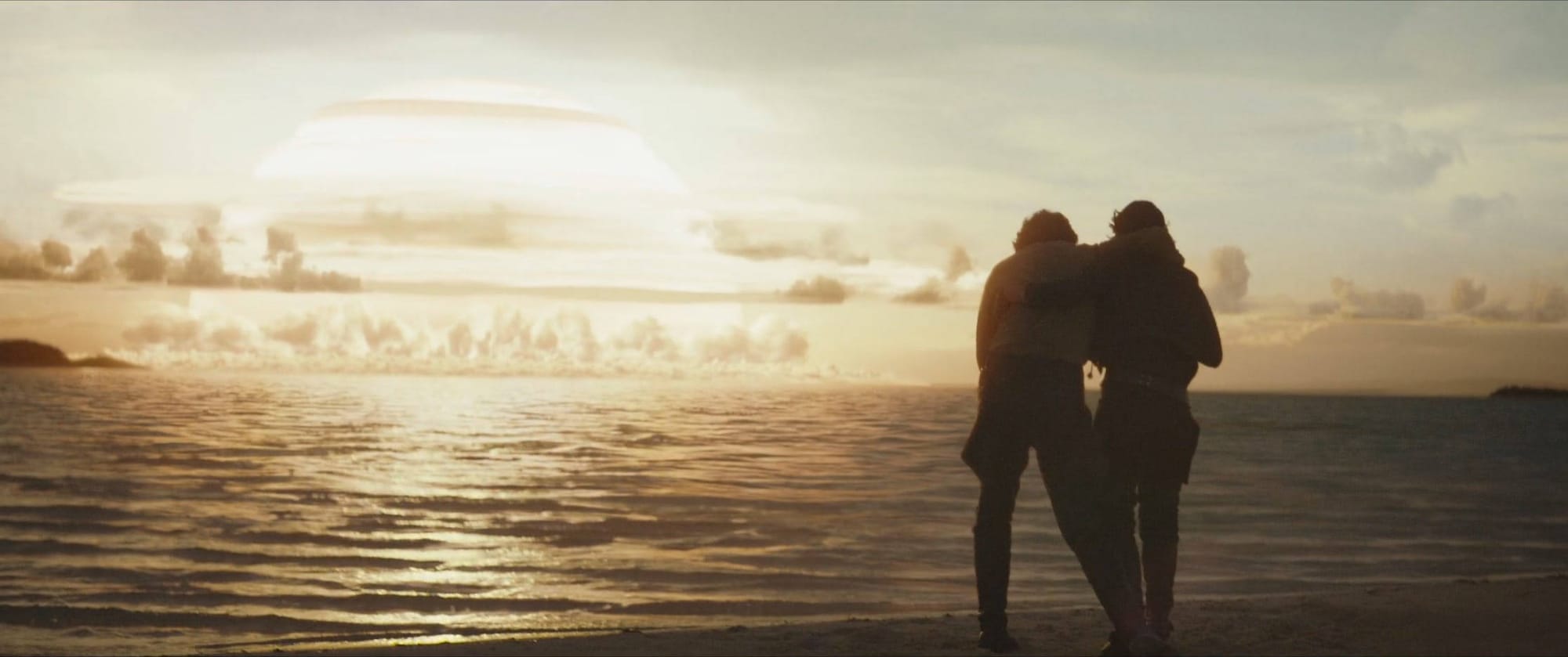 Cassian and Jyn look out into the water on a beach. An explosion nearby is racing across the water towards them.
