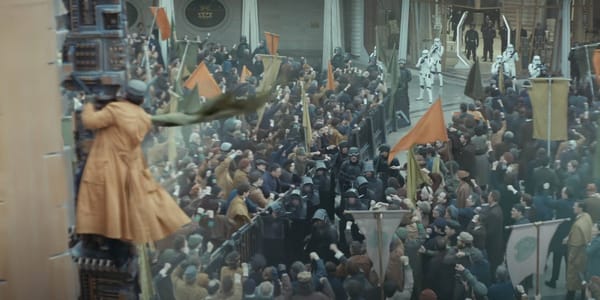 A crowd of protesters circle riot cops who are seperated by a barrier. A protester waves a flag while hanging from a pole in the foreground. Storm Troopers are in the background.